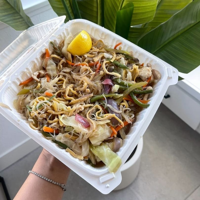 A hand holds a takeout container of Filipino pancit with noodles, vegetables, and a lemon wedge.