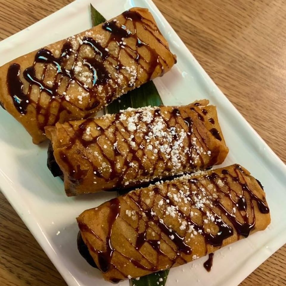 Three Filipino turon desserts, drizzled with chocolate sauce and powdered sugar, on a white plate.
