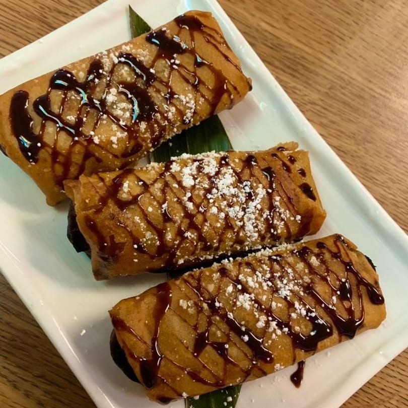 Three Filipino turon desserts, drizzled with chocolate sauce and powdered sugar, on a white plate.