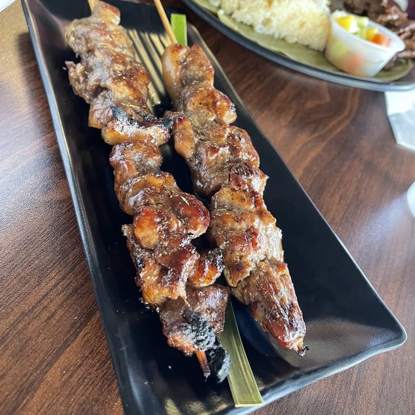 Two skewers of grilled Filipino pork barbecue, glistening with sauce, on a black rectangular plate.